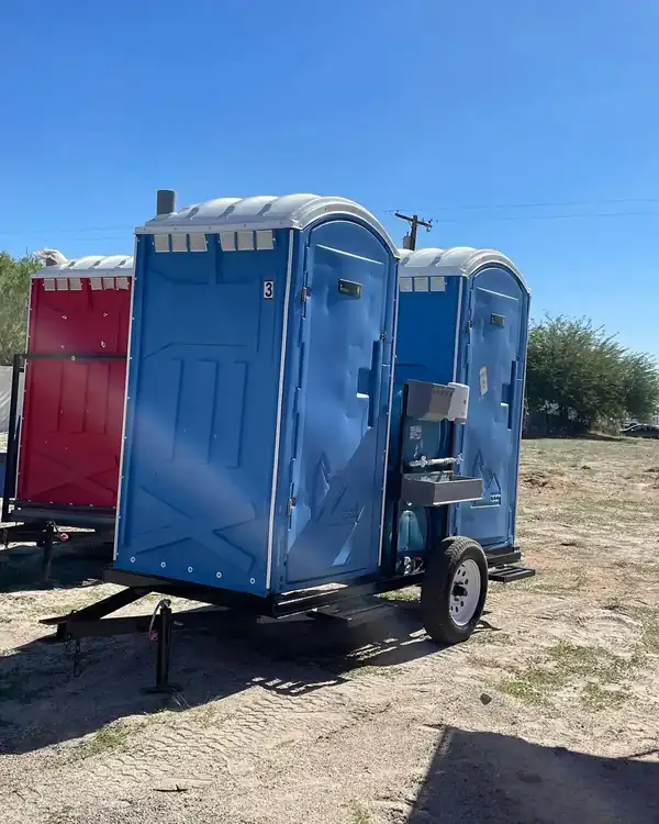 Blue porta potty mounted on a small towable trailer at a dirt worksite